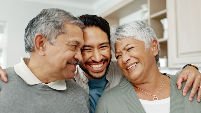 A young adult man hugs his parents. All are smiling, as he's learned how to talk to your parents about money. Image by Yuri Arcurs