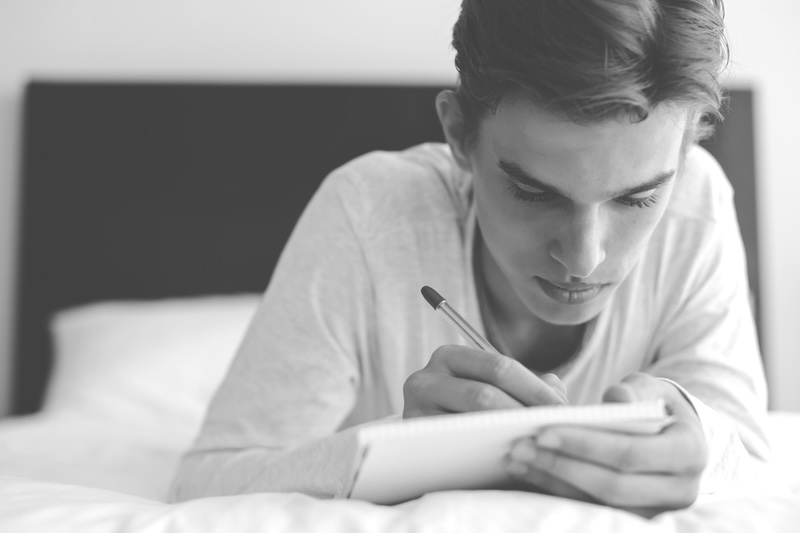 A young man journaling for self-awareness, lying on his bed. By Alena Ozerova