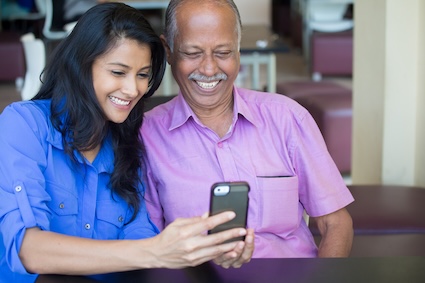 A granddaughter and grandfather looking at smartphone and smiling. By Nanditha Rao. Article on the best cell phone for seniors