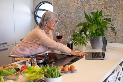 Woman in her kitchen preparing healthy meals for seniors who live alone while drinking wine and using her laptop. Image by Lucigerma