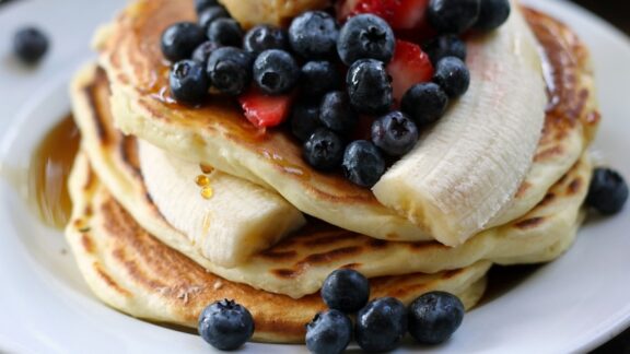 high-protein pancakes with blueberries and bananas and syrup on them.
