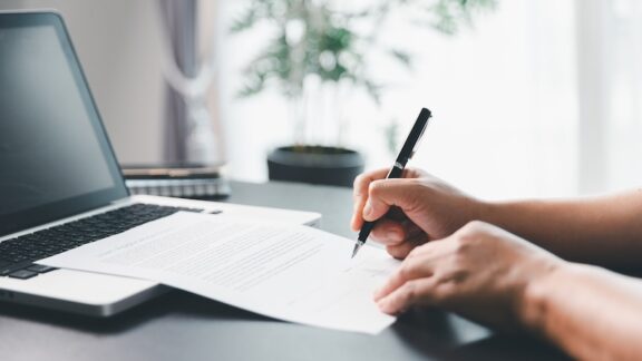 Senior writing on paper in front of computer getting their estate planning in order.