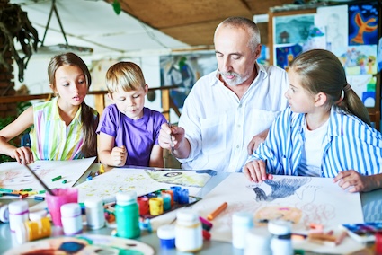 Senior man teaching art to three children, modeling the benefits of intergenerational programs. Image by Seventyfourimages