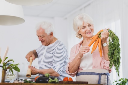 A woman holds up carrots in her kitchen as she and her partner prepare a meal using long-lasting vegetables. Image by Bialasiewicz.