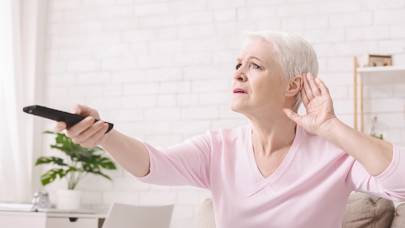 A woman holding her hand to her ear and pointing the remote at a TV, indicating she has hearing problems. Hearing care is brain care.