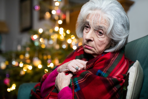 A woman sad in a festive blanket in front of a holiday tree going through Christmas disappointment.
