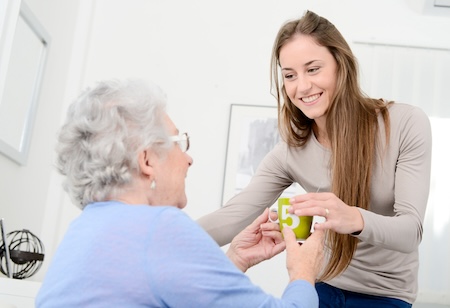 Woman visiting an older woman at home, bringing her a beverage. By JPWallet. For article on online care coordination platforms.