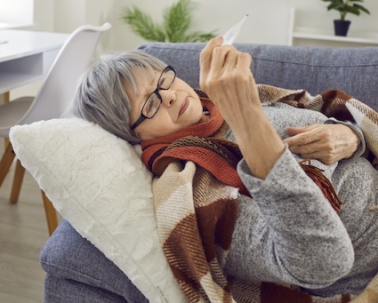 A sick woman lying on a sofa, wrapped in a blanket and checking her temperature on a thermometer, not having known how to stay safe in flu season. By Lacheev
