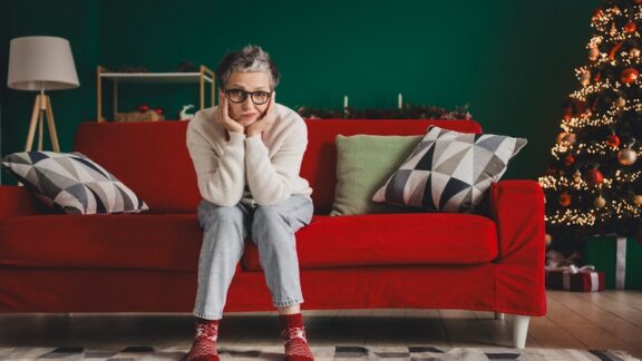 A woman sitting on her couch wondering about managing holiday stress, with the Christmas tree behind her.