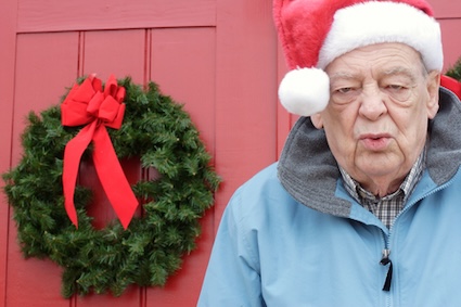 A man with a Santa hat on and a Christmas wreath behind him, trying to focus on managing holiday stress. By Donnarae