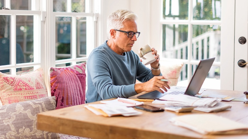 A man at home on his laptop, surrounded by papers. Seniors can educate themselves and use critical thinking skills for safer online experiences.