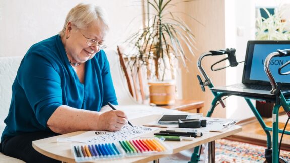 A woman taking an online art class, which also helps her accentuate the positive with joy and creativity; with her computer perched on her rollator and colored pens on her desk. By Irinayeryomina