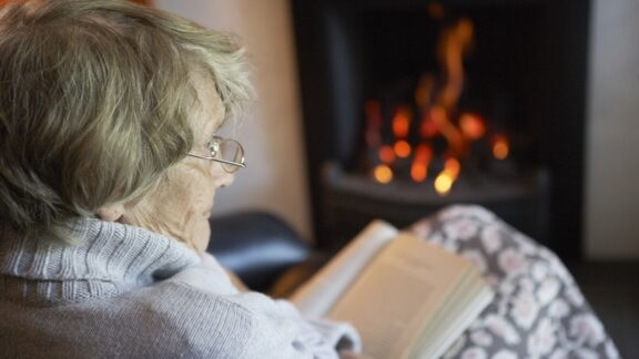 A woman wrapped up warm in front of a fireplace and reading, managing chronic conditions in cold weather