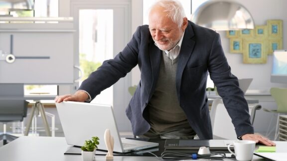 A senior man in his office looking at his laptop, after delaying retirement. Nyul