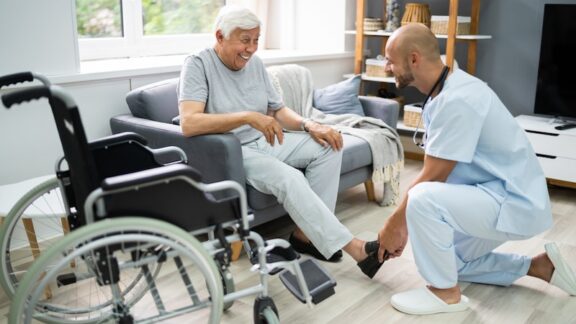 Old Senior Home Care. Nurse Helping Patient To Dress Shoes, for decision between aging in place or assisted living