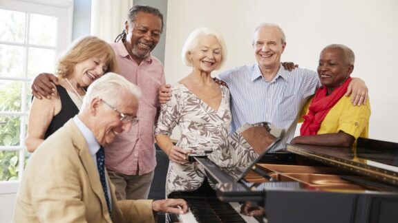 A group of older adult friends gathered around a computer as one man plays. They have determined how to pay for a senior living community. Monkey Business Images