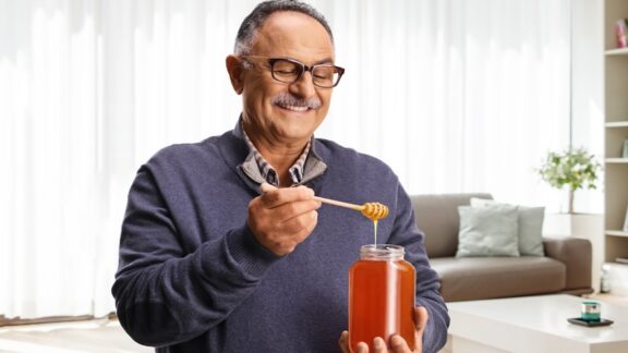 A mustached senior smiling at a jar of honey, because honey can enhance your mood. Ljupco