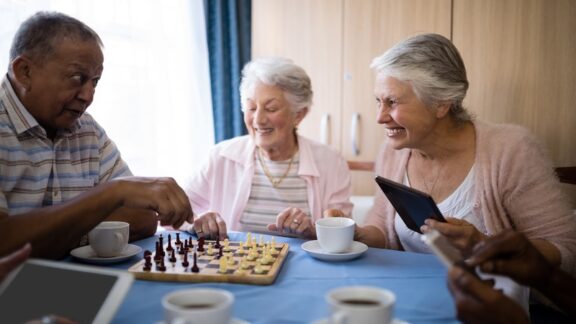 Friends playing chess in an independent living community common area, having learned the benefits as they accept help. Wavebreakmedia Ltd
