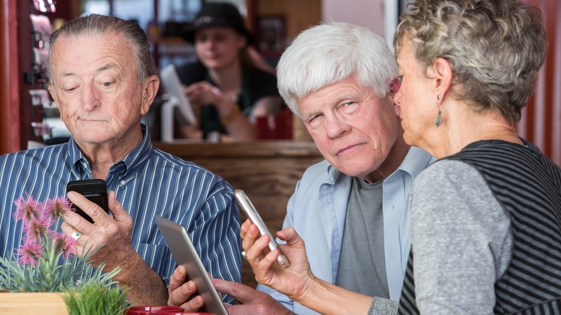 senior friends in a coffee shop, all looking medical conditions up on their smart devices. Is it OK to talk about our health? Creatista