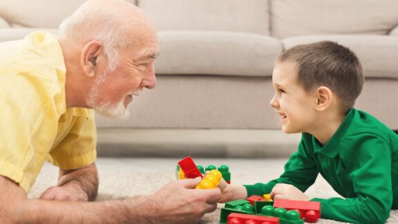 Senior man and young boy playing large Lego-style blocks at intergenerational care centers. Prostockstudio