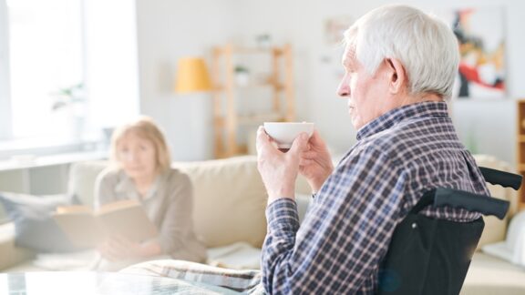 Caregiving can be stressful. A man in a wheelchair sips his coffee while his wife sits in the background. Caregiver respite programs can help. Pressmaster